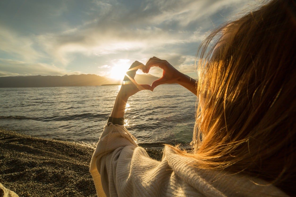 A woman standing on the beach, smiling, making a heart shape with her hands against the backdrop of the ocean.