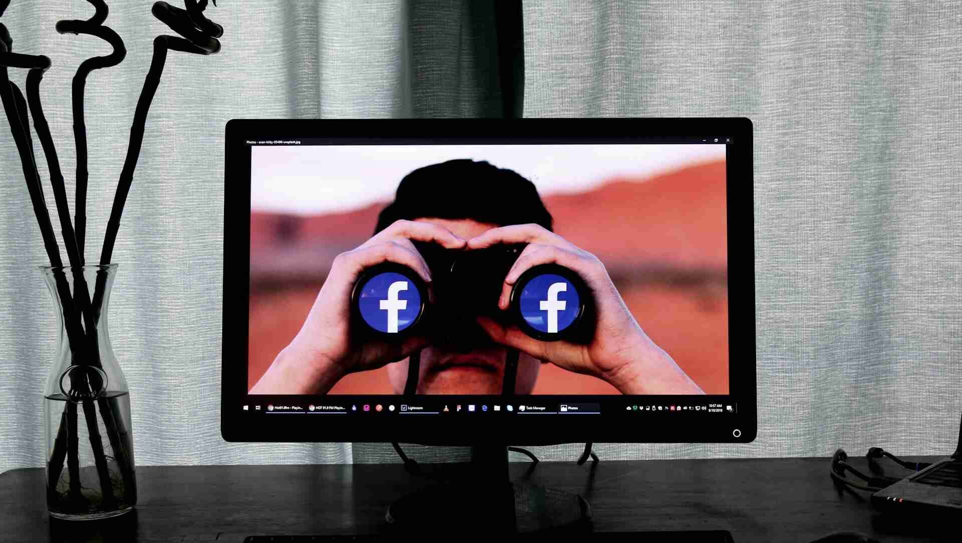 Person seen on a computer screen looking through binoculars with Facebook logos, symbolising social media obsession and the addictive nature of constant Facebook use.