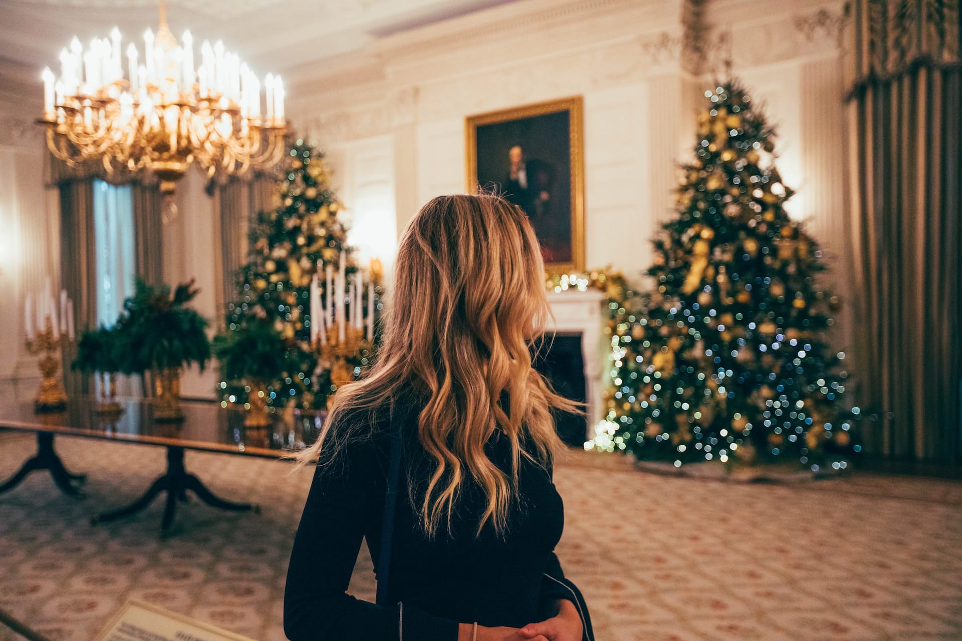 A woman sits in a festive living room, looking out the window with a thoughtful expression, feeling lonely